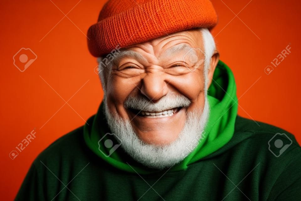 Cierra el retrato de un feliz hombre optimista de 70 años con la cara arrugada sonriente, vestido con un sombrero naranja hipster y una capucha verde, aislado sobre fondo negro. Positivo y alegre a cualquier edad.