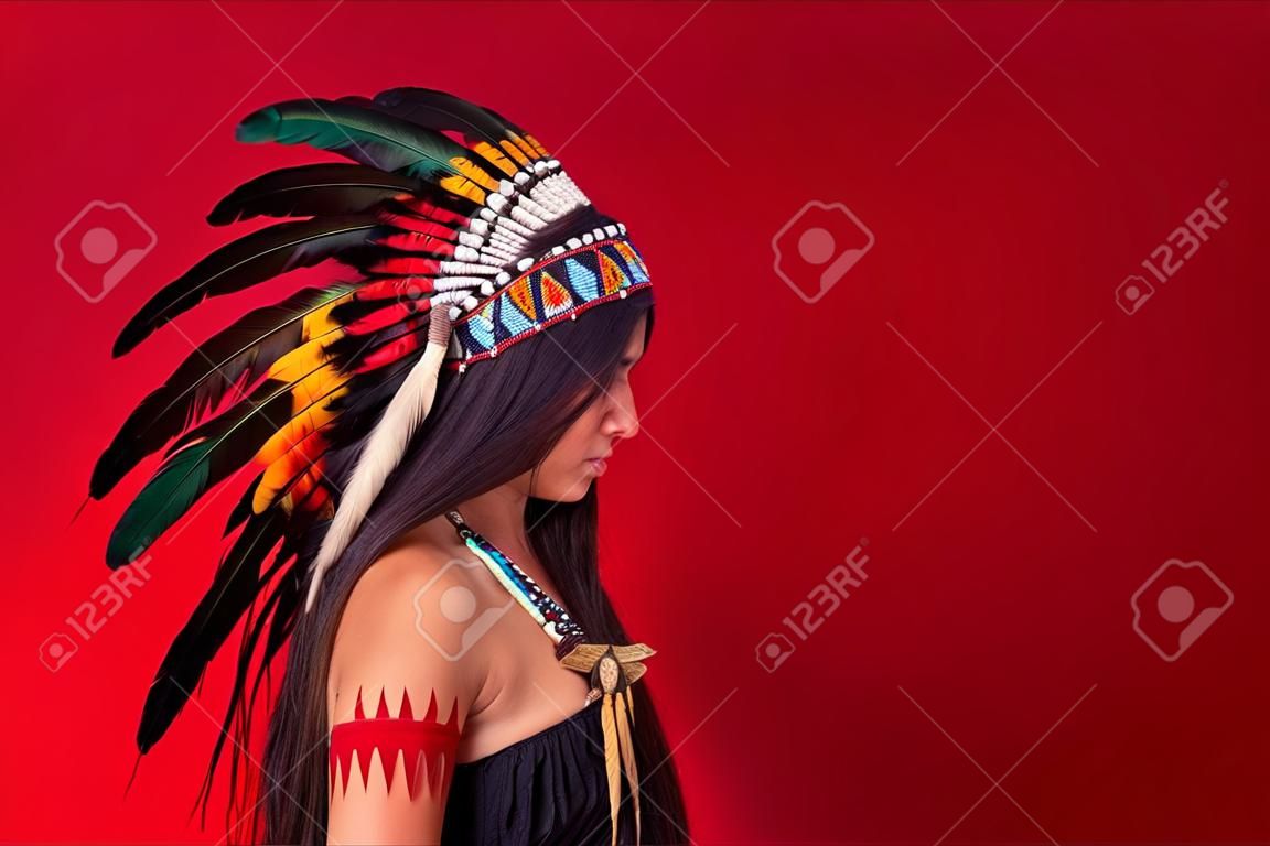vista laterale sulla donna sciamana dai capelli lunghi con piume sulla testa che guarda in basso, costume indiano aborigeno, isolato su sfondo rosso. etnia, individualità