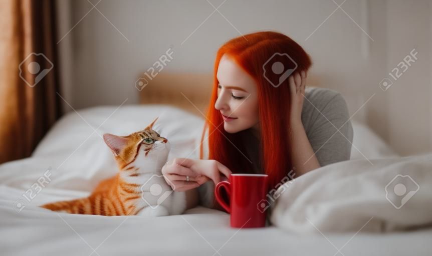 Retrato de uma mulher atraente, contente, jovem, sexy, cabelo longo vermelho brilhante tingido, deitado relaxado na cama da manhã goza e abraça com seu gato, com café em um copo vermelho, espaço da cópia