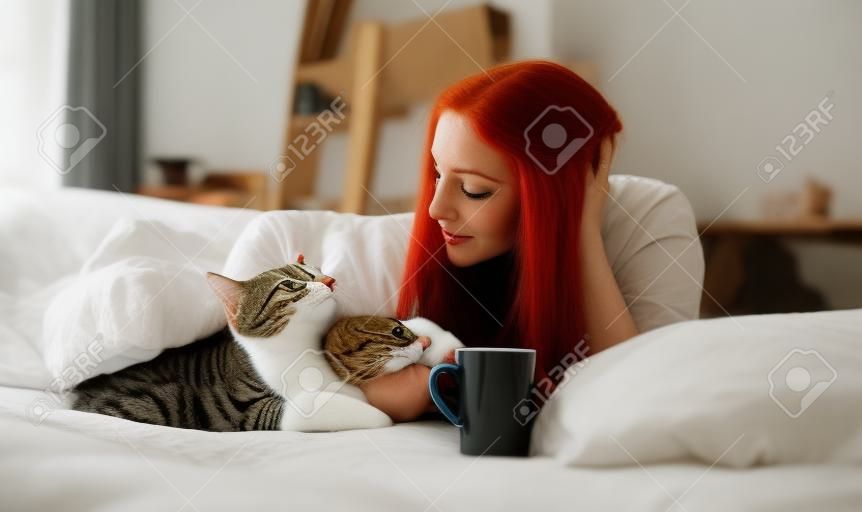 Retrato de uma mulher atraente, contente, jovem, sexy, cabelo longo vermelho brilhante tingido, deitado relaxado na cama da manhã goza e abraça com seu gato, com café em um copo vermelho, espaço da cópia