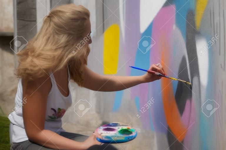 mulher artista feminina morena em camisa branca sexy, na luz solar quente da noite, pintura com pincel e paleta na frente de um grande mural, pinta uma borboleta de pavão na parede, espaço de cópia