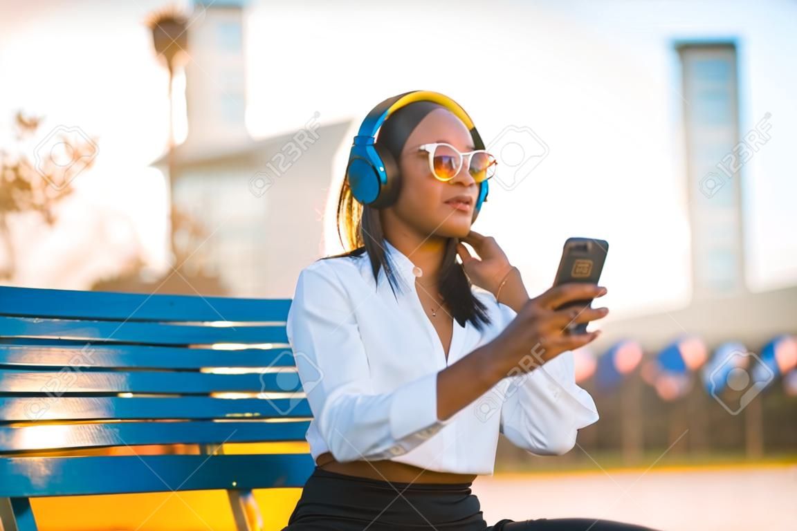 African american woman listening to music and smiling with headphones in the summer at sunset in the city