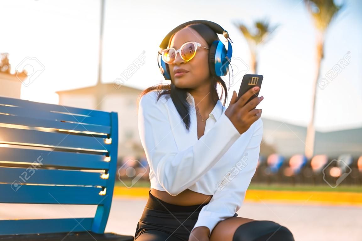 Mujer afroamericana escuchando música y sonriendo con auriculares en el verano al atardecer en la ciudad