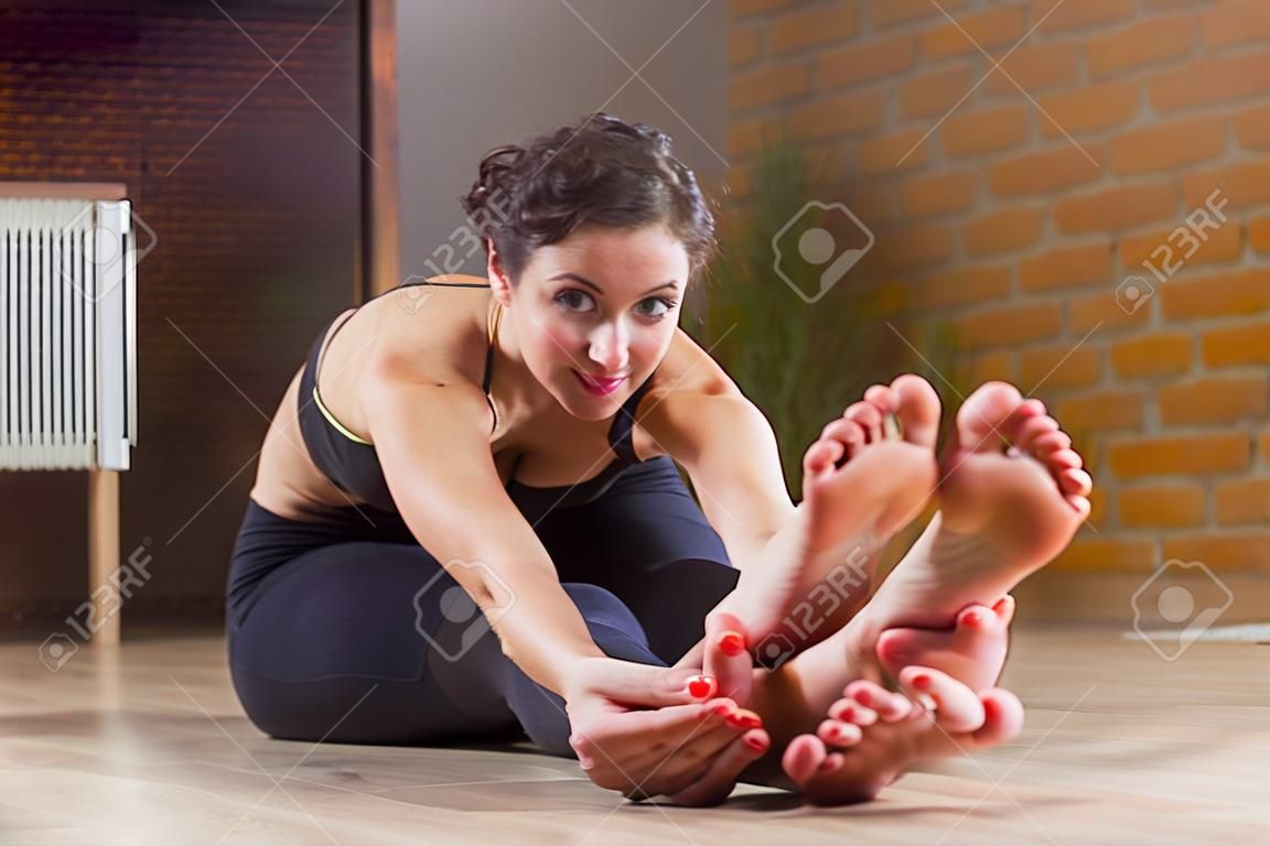 Pretty European woman sitting barefoot stretching her back and legs  on floor bending forward looking at camera