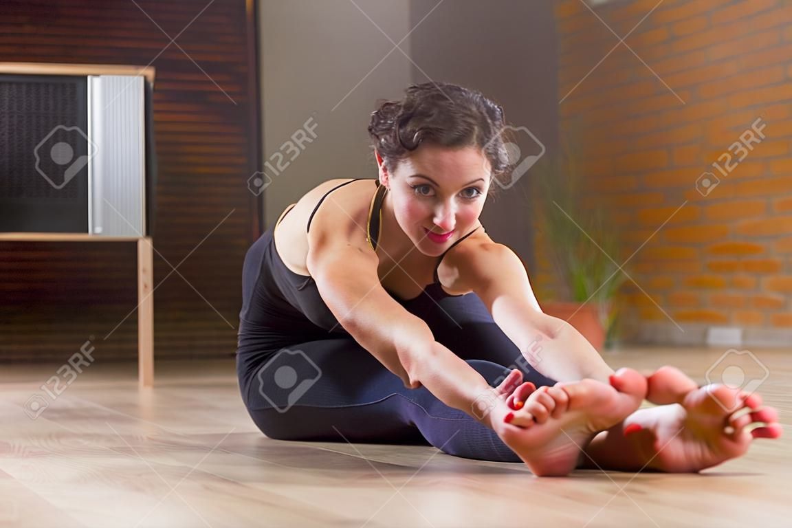 Pretty Europejskiej kobieta siedzi boso podciągania jej pleców i nóg na podłodze gięte do przodu spojrzenie na aparat fotograficzny