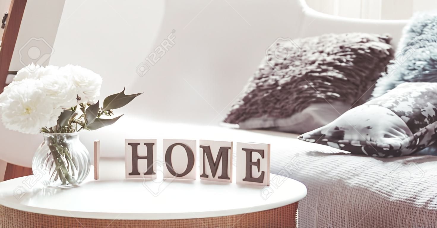 Chic interior for a house. Candles, a vase with flowers with wooden letters of the home on wooden white chair. Sofa and wicker basket with cushions in the background. Home decoration.