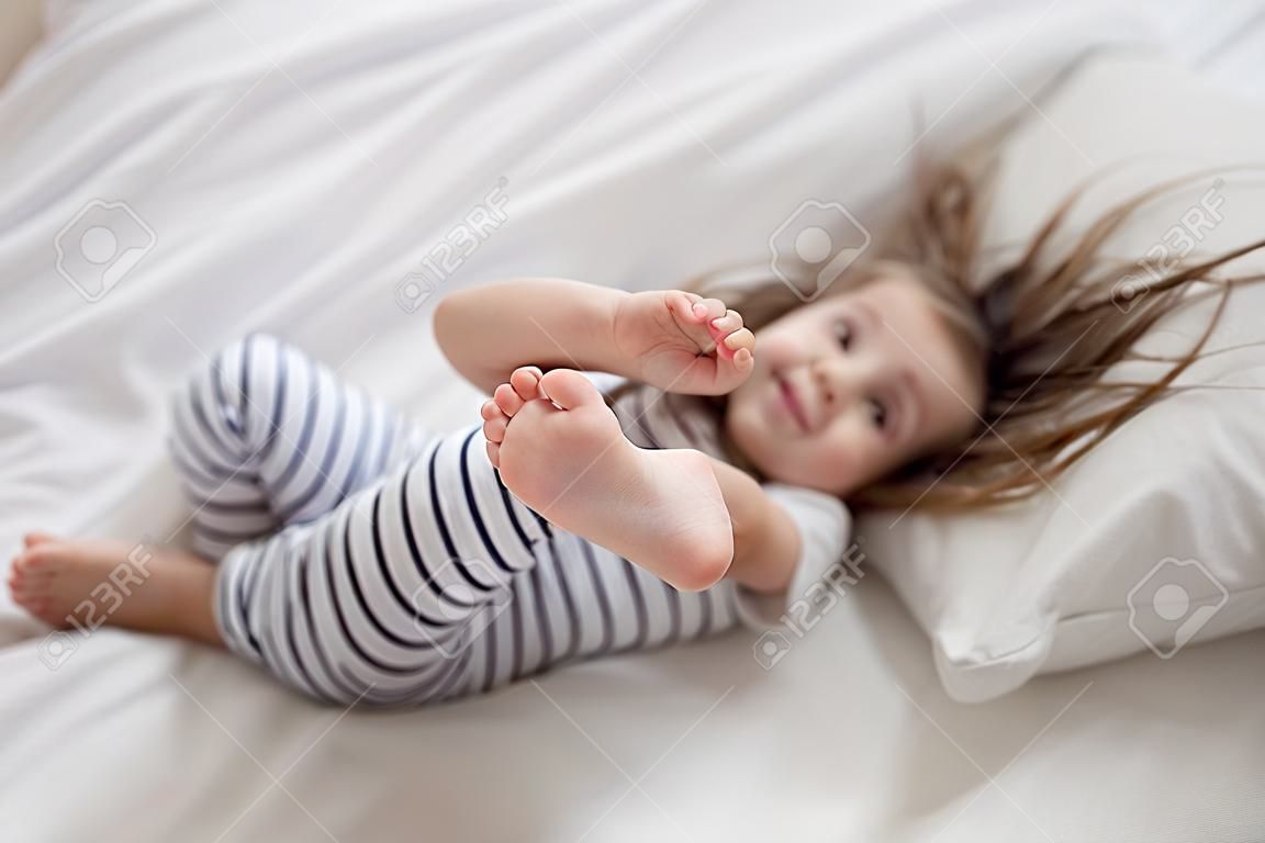 Cute little fun girl with long hair in bed woke up in the morning . Concept of sleep and development of children.
