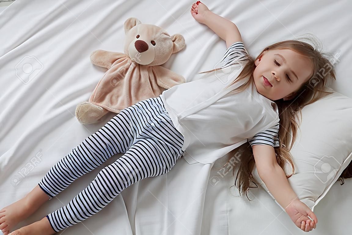 Nettes kleines Mädchen im weißen Bett, das mit weichem Bärenspielzeug spielt. Konzept des Schlafes und der Entwicklung von Kindern.