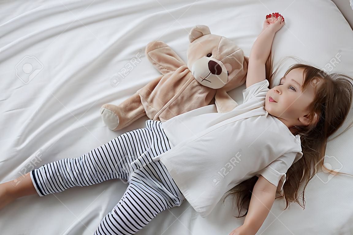 Nettes kleines Mädchen im weißen Bett, das mit weichem Bärenspielzeug spielt. Konzept des Schlafes und der Entwicklung von Kindern.