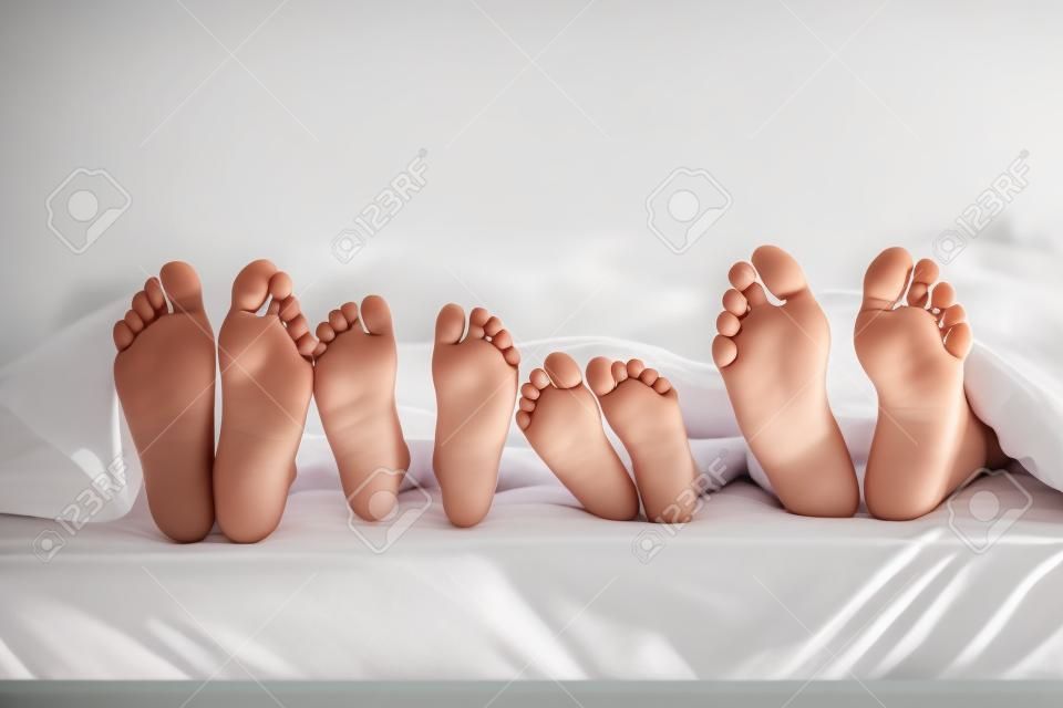 Madre, padre y dos niños se encuentran en la cama con sábanas blancas; centran en los pies