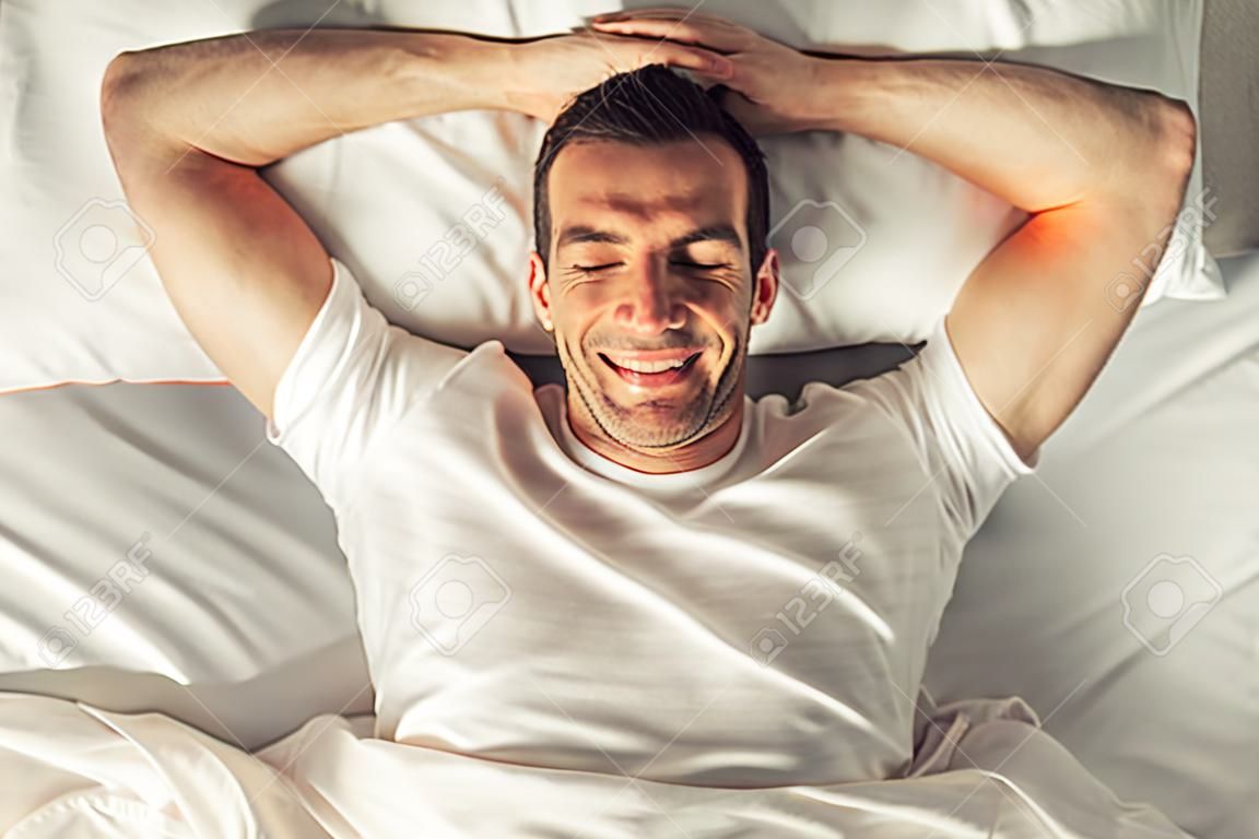 Top view of handsome man smiling while lying with closed eyes and hands behind head in his bed at home