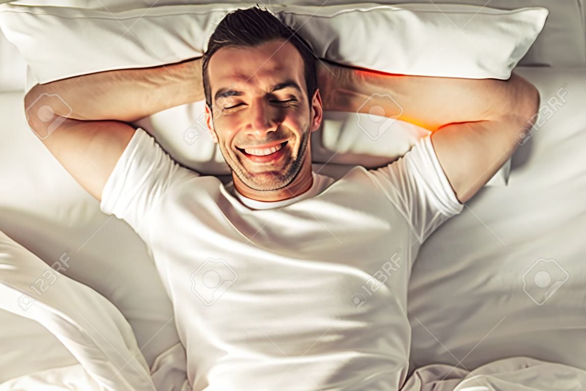 Top view of handsome man smiling while lying with closed eyes and hands behind head in his bed at home