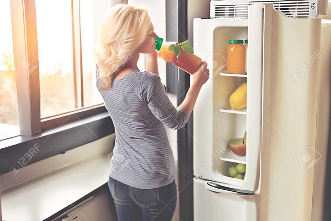 Güzel kız bir sürahi meyve suyu tutuyor ve evde mutfakta açık buzdolabının yanında dururken gülümsüyor