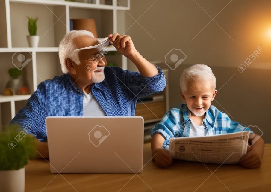 Дедушка и внук отдыхают дома. Красивый старик использует ноутбук, пока его мальчик читает газету