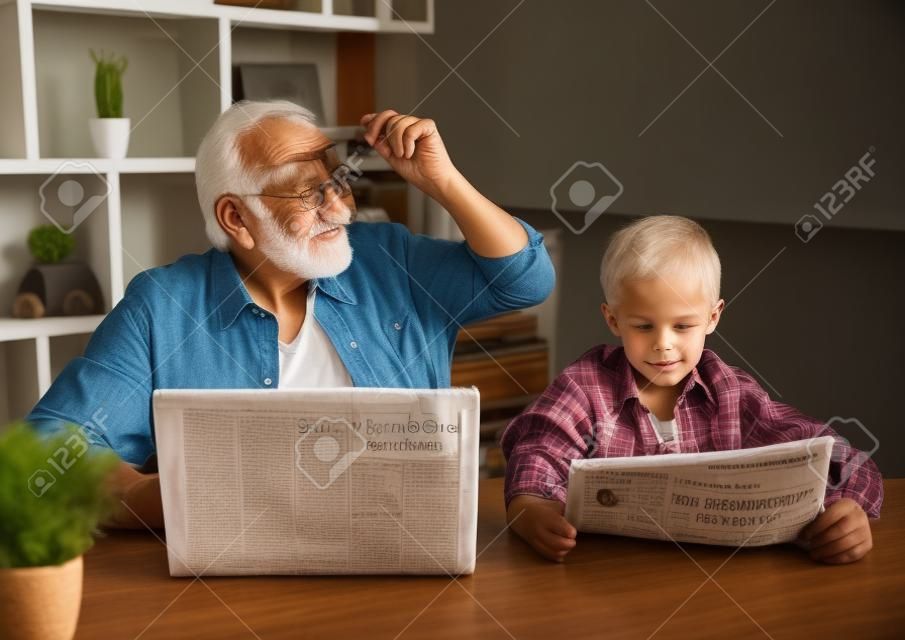 Opa en kleinzoon rusten thuis. Knappe oude man gebruikt een laptop terwijl zijn jongen een krant leest