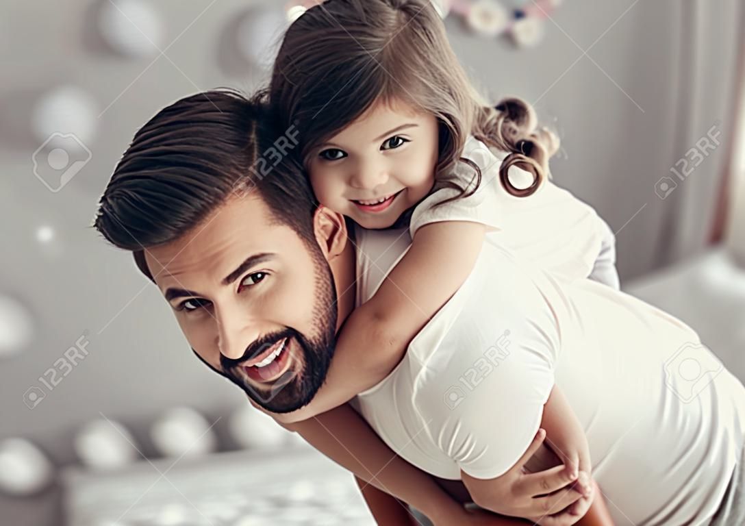 Der hübsche junge Vater und seine niedliche kleine Tochter spielen zusammen im Kinderzimmer. Mädchen sitzt Pickaback