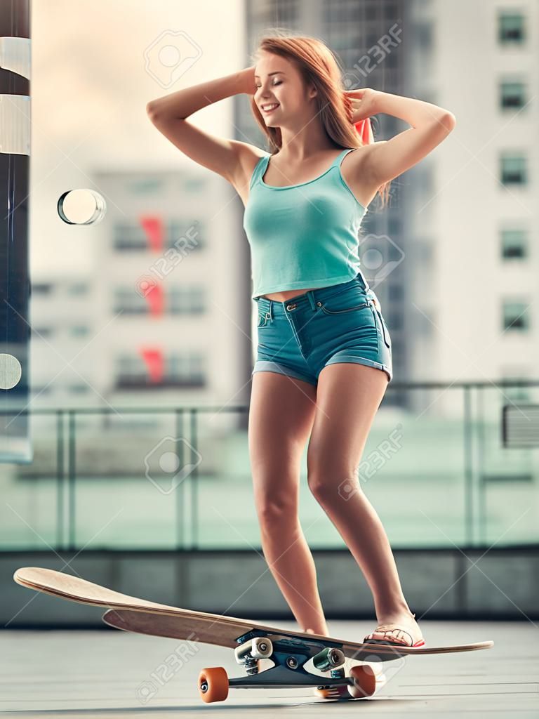 Stilvolles junges Mädchen lächelt beim auf Skateboard draußen fahren