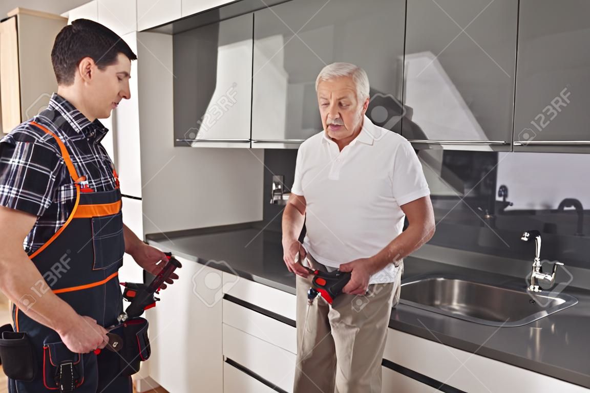 Due uomini di idraulici sono in piedi in cucina. Un uomo anziano ispeziona il luogo del lavoro di riparazione.