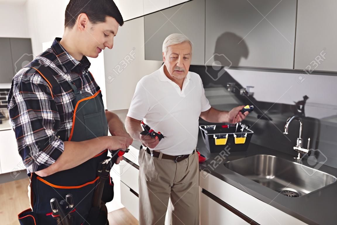 Due uomini di idraulici sono in piedi in cucina. Un uomo anziano ispeziona il luogo del lavoro di riparazione.