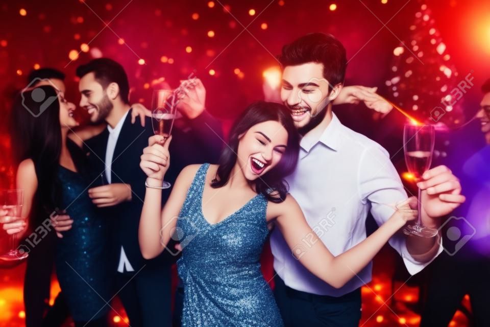 Young people have fun at a New Years party. In the foreground, a couple is dancing with glasses in their hands.