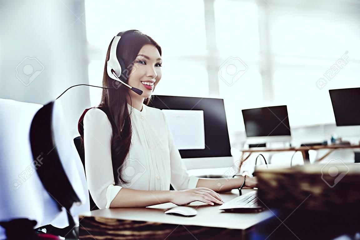 Chica asiática sentada en el call center. Ella tiene auriculares en los que habla con los clientes. Hay una computadora frente a ella.