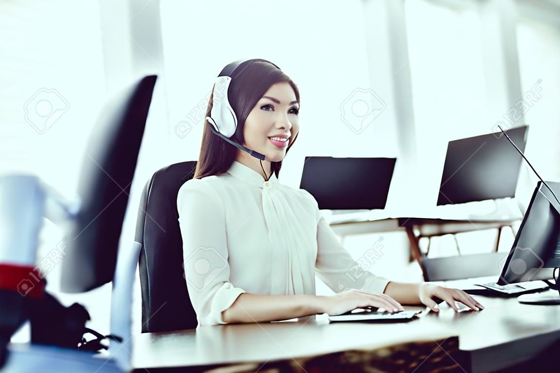Asian girl sitting in the call center. She has headphones on which she talks to customers. There is a computer in front of her.