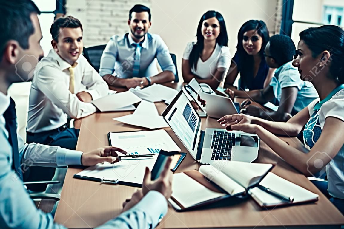 Young Smiling Business People on Meeting in Office. Group of Young Coworkers Sitting Together at Table in Modern Office and Listening to Businessman Presenting. Teamwork Concept. Corporate Lifestyle