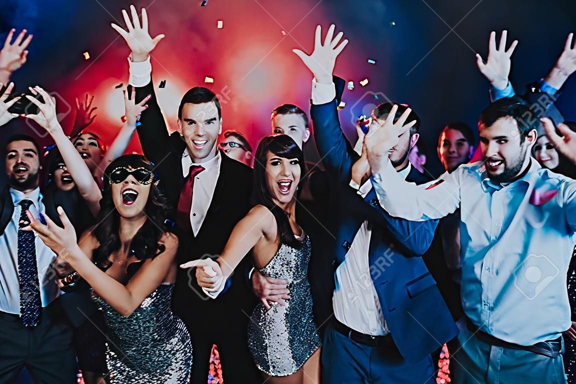 Smiling People Celebrating New Year on Party. Happy New Year. People Have Fun. Indoor Party. Celebrating of New Year. Young Woman in Dress. Young Man in Suit. Excited People. Hands Up.