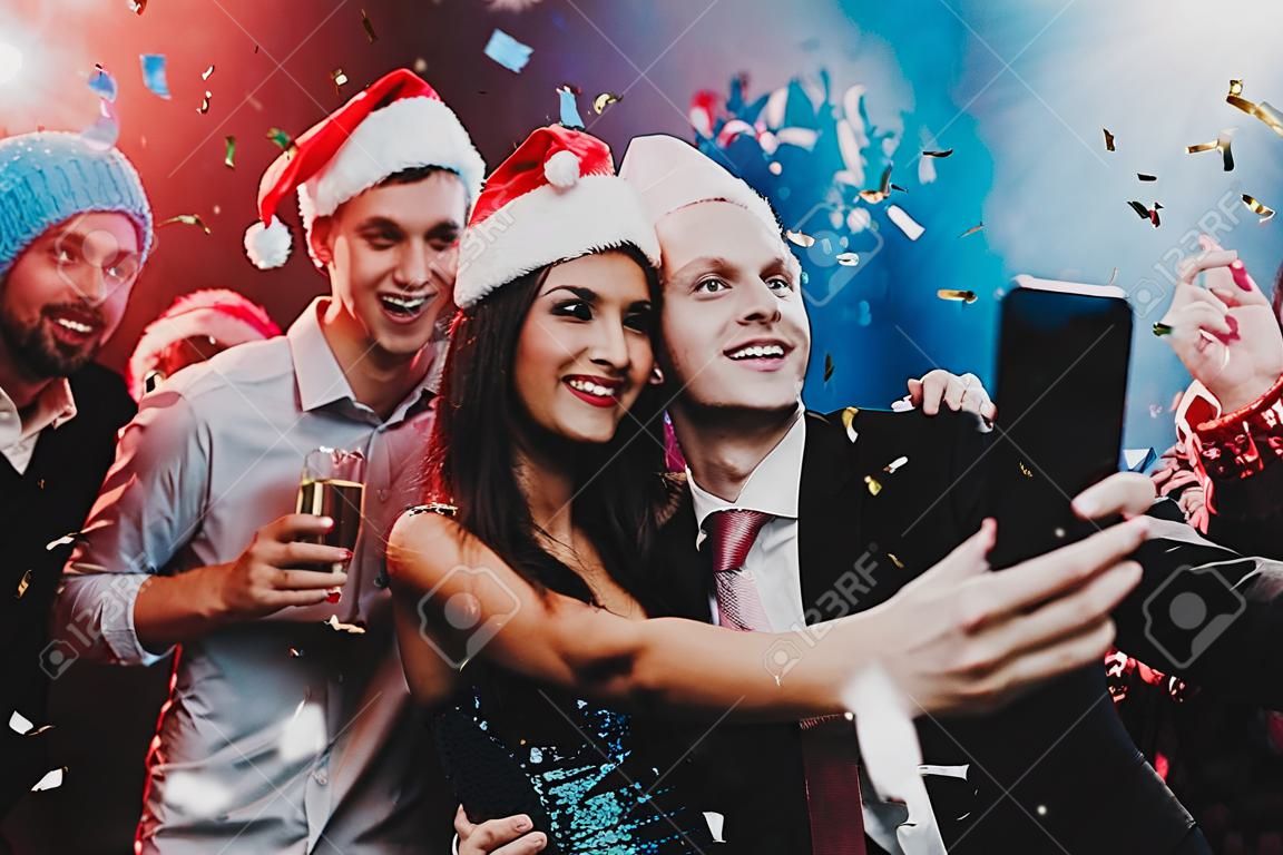 Happy People Taking Selfie on New Year Party. Happy New Year Concept. People Have Fun. Indoor Party. Celebrating of New Year. Young Woman in Dress. Young Man in Red Cap. Using Smartphone.