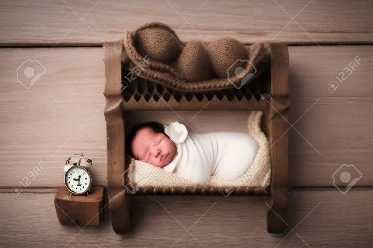 Tiny newborn twin boys in white bodysuits against a light wood background. Newborn twins sleep on a bunk wooden bed. The boys are dressed in white caps. Clock with time of birth. Studio photography.
