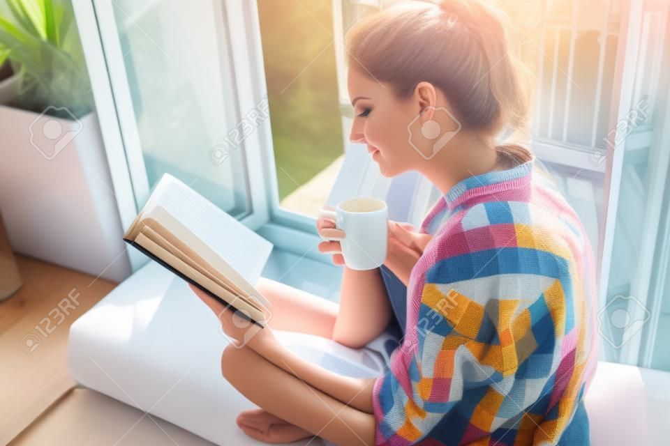 젊은 예쁜 여자 열린 창에 앉아 커피를 마시고 책을 읽고는 나머지 즐긴다