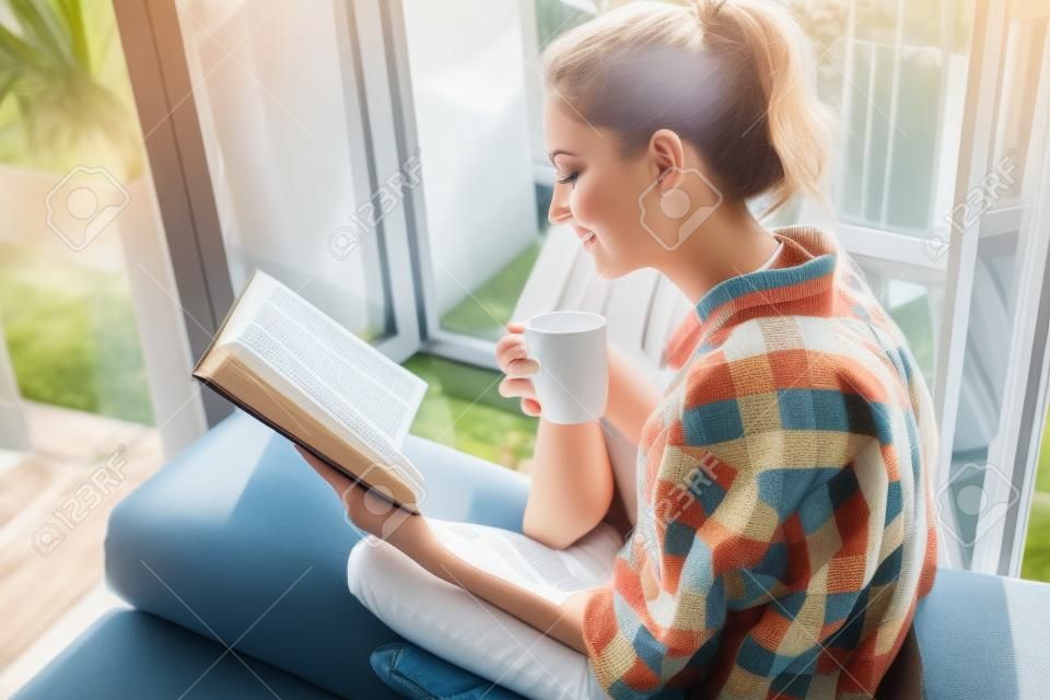 Jovem mulher bonita sentado na janela aberta bebendo café e lendo um livro goza de descanso