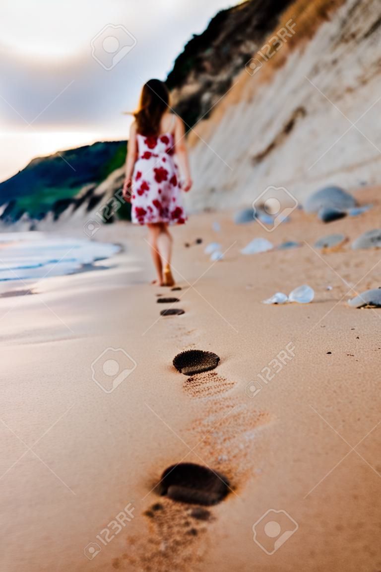 Beauty young woman in stylish dress walking barefoot by the beach leaving footprints in sand at sunset over the horizon. Travel and vacation concept.