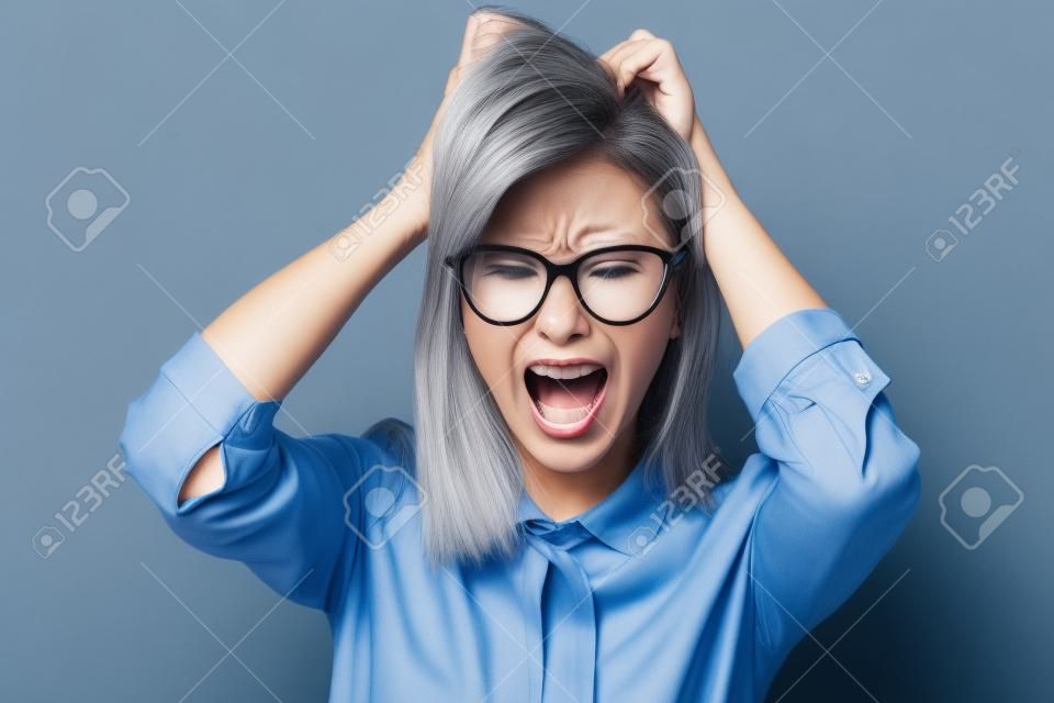 Angry businesswoman toccando i capelli e urlando su sfondo grigio. Indossare in camicia blu e occhiali. Guardando la fotocamera