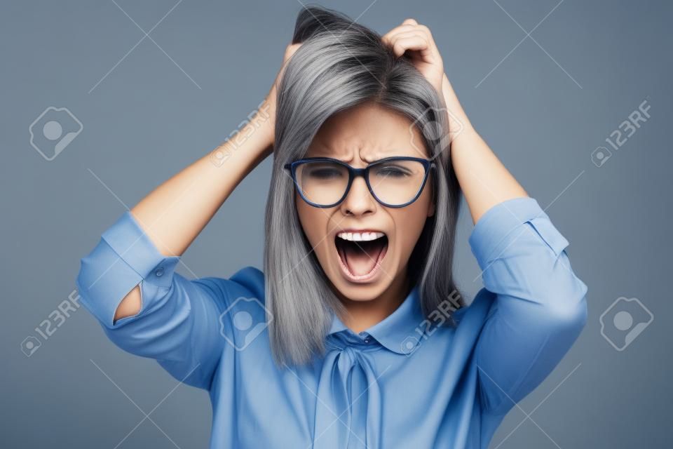 Boze zakenvrouw aanraken haar haar en schreeuwen over grijze achtergrond. Draagt in blauw shirt en bril. Kijken naar camera