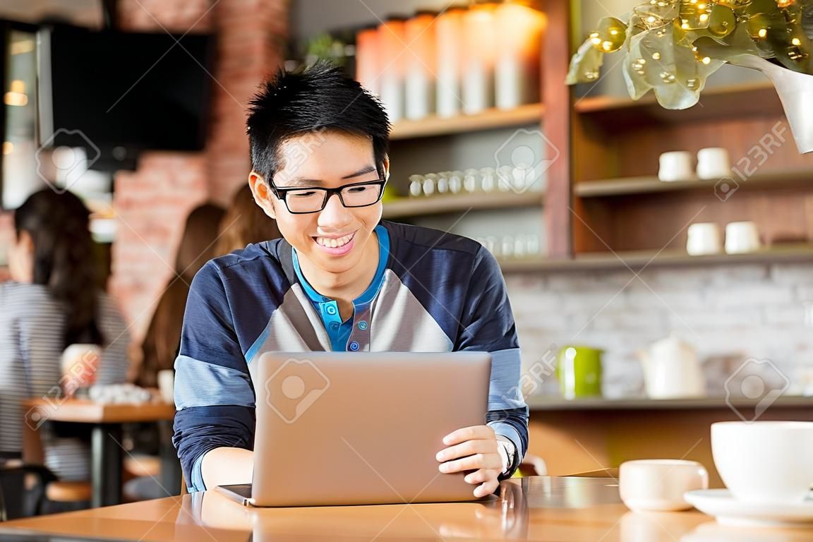 Happy cheerful young asian male in glasses smiling and using laptop in cafe