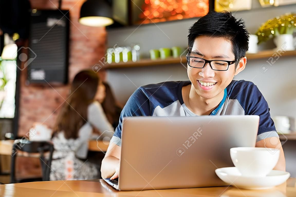 Felice allegro giovane maschio asiatico in bicchieri sorridente e con laptop in caffè