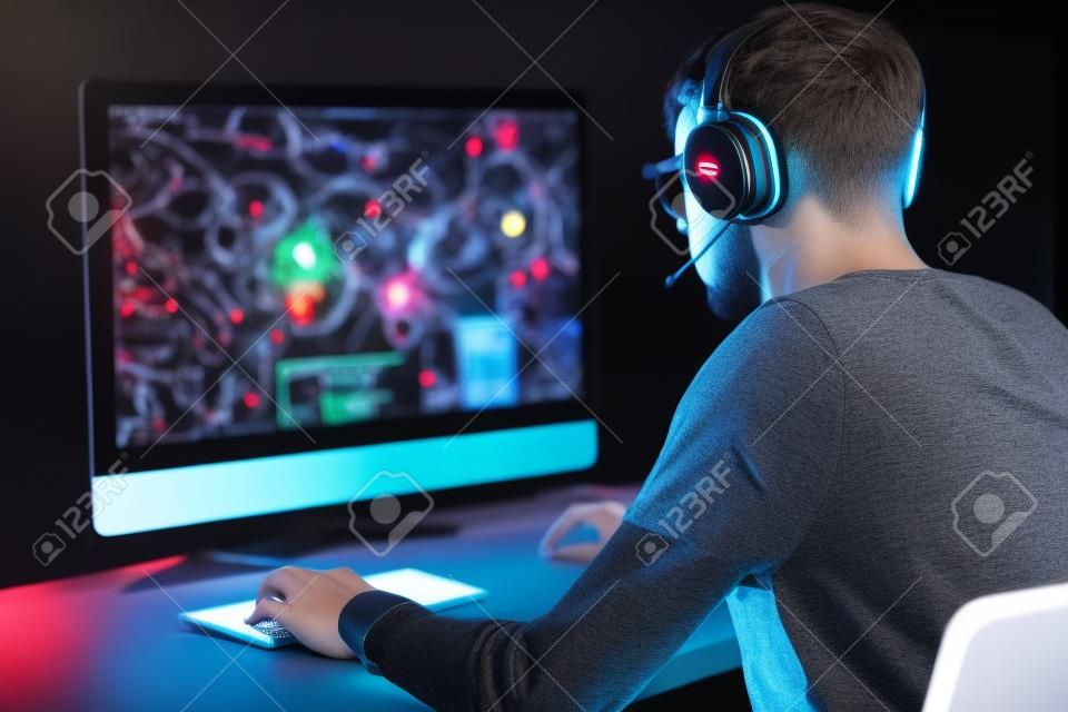 Back view of concentrated young gamer in headphones and glasses using computer for playing game at home