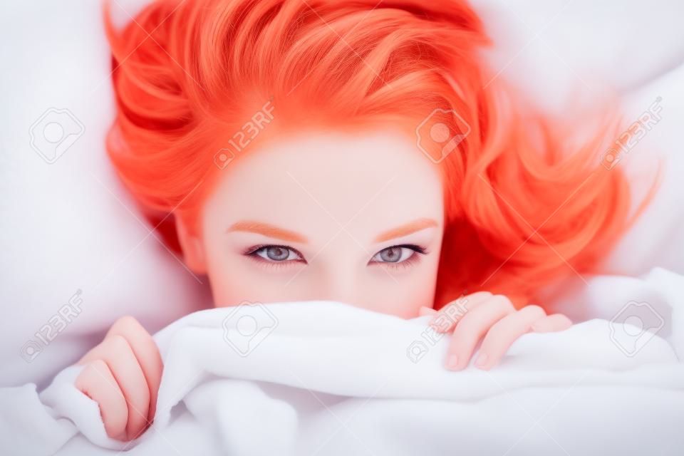 Closeup of cute lovely young woman with red hair lying and hiding under white blanket