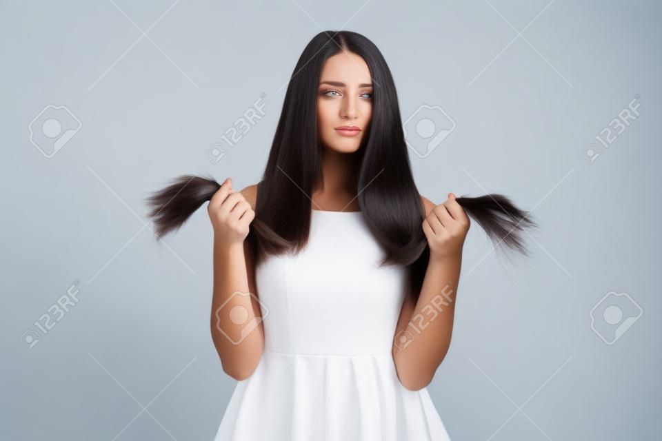 Upset beautiful young woman standing and looking on splitting ends of her long dark hair over white background