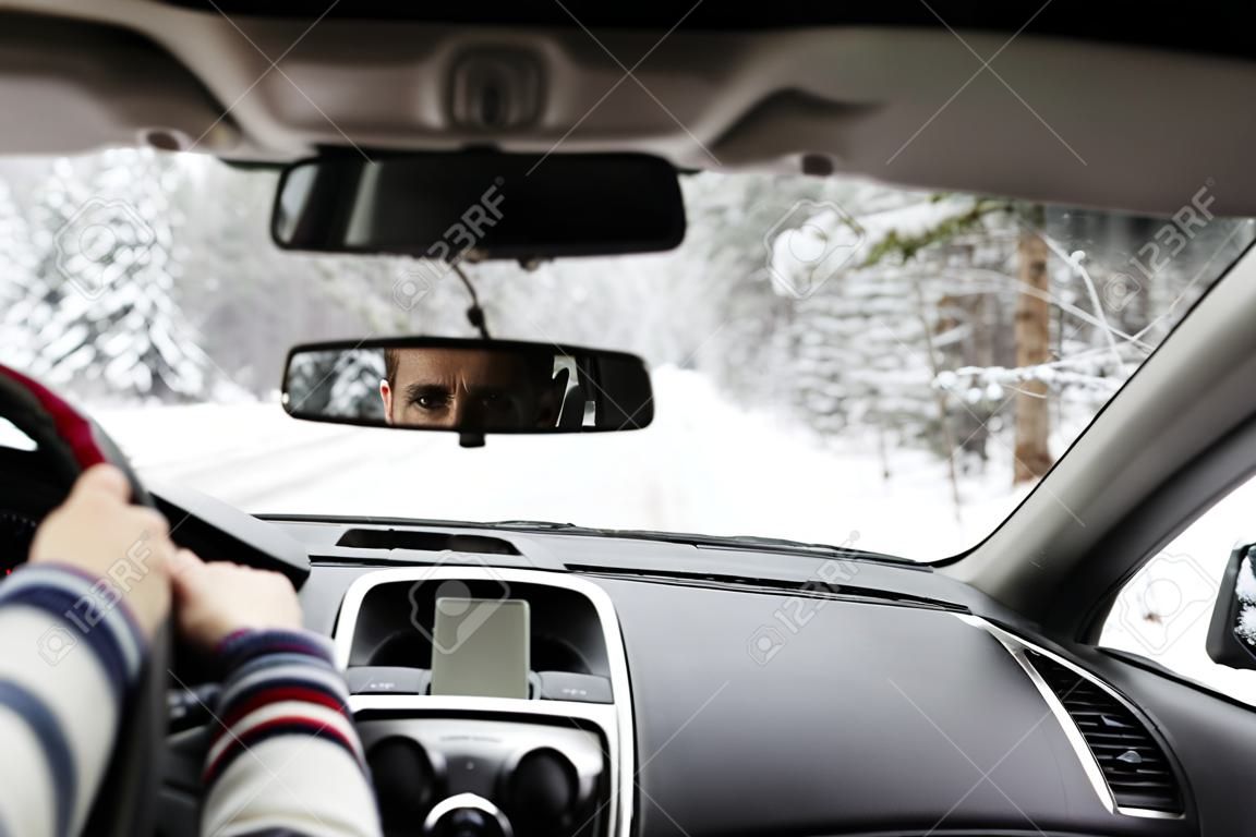 Closeup of eyes of young man reflected in car rear viewer while driving in winter forest