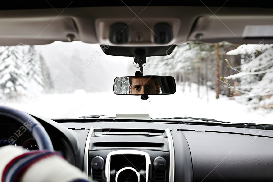 Il primo piano degli occhi del giovane ha riflesso nel visore posteriore dell'automobile mentre guidava nella foresta dell'inverno