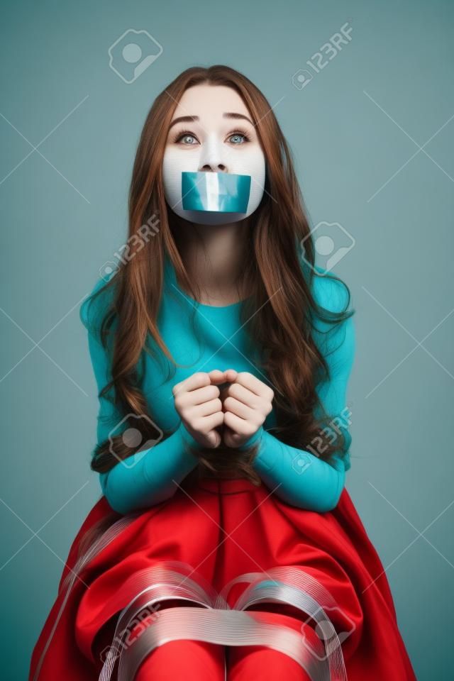 Frightened young woman bounded by ropes sitting with closed mouth by tape