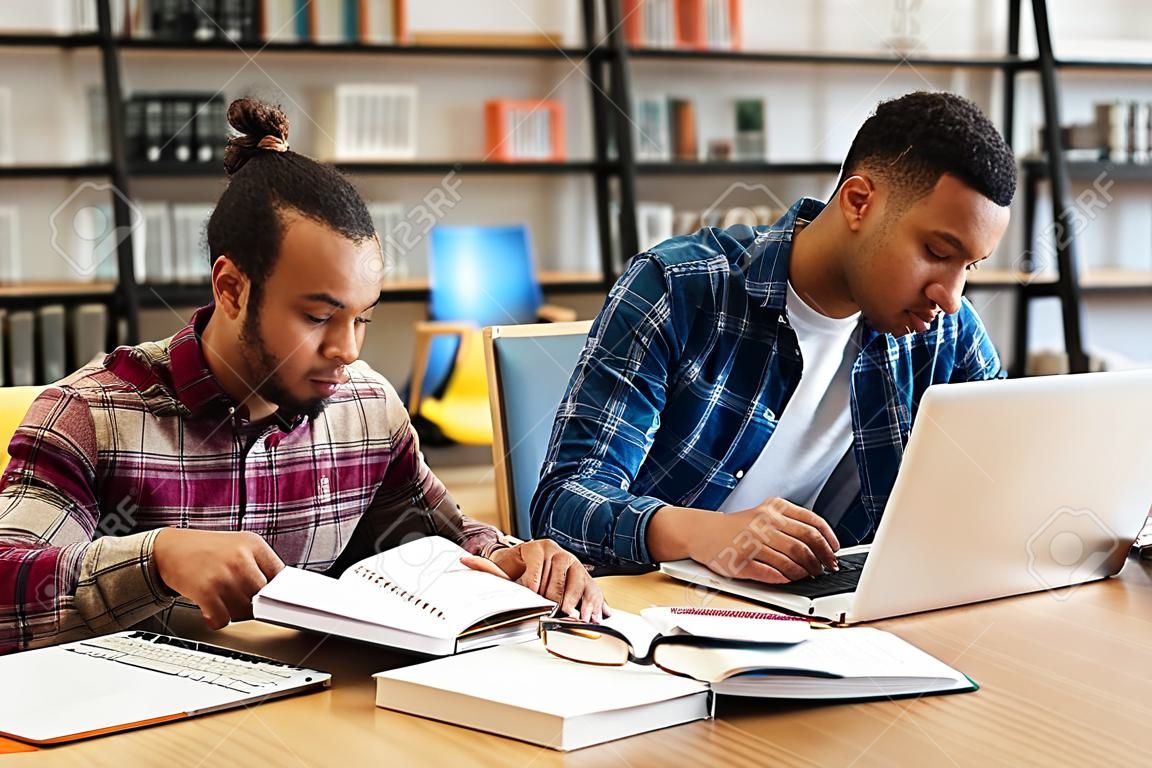 Twee gemengde race mannelijke studenten studeren met laptop terwijl zittend aan de bibliotheekbalie