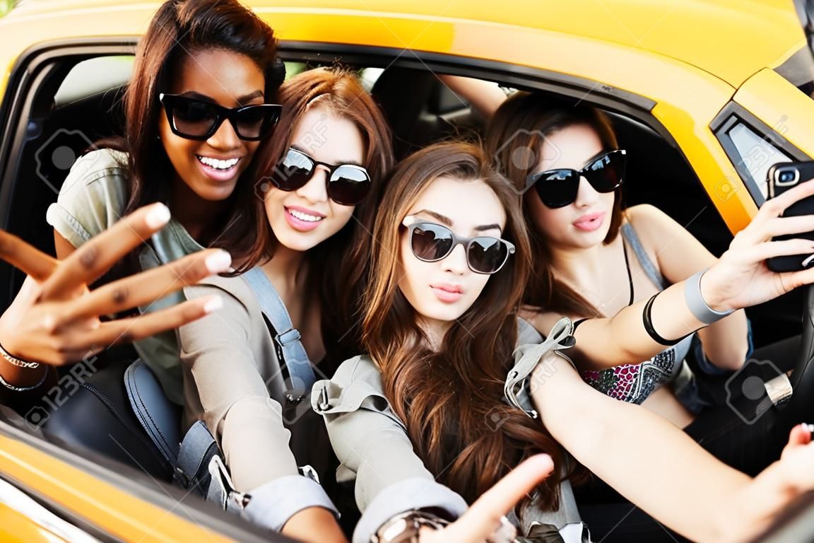 Picture of smiling emotional four young women friends sitting in car outdoors. Make selfie by phone.