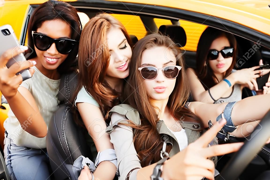 Zdjęcie uśmiechniętych, emocjonalnych czterech młodych przyjaciółek siedzących w samochodzie na zewnątrz, robiących selfie telefonem