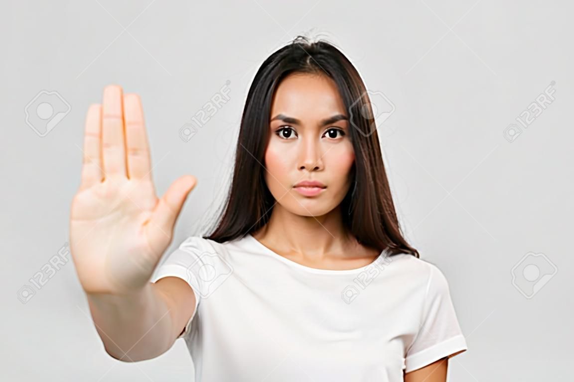 Ritratto di una donna asiatica giovane seria che sta con il gesto steso di arresto di rappresentazione della mano isolato sopra fondo bianco