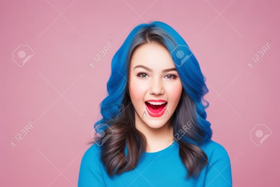 Chiuda sul ritratto di una ragazza allegra vestita in maglione che esamina la macchina fotografica e sbattere le palpebre isolato sopra fondo blu
