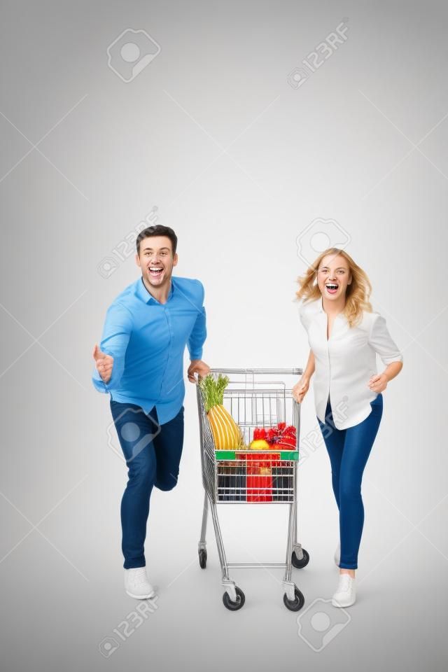 白い背景の上に隔離されたスーパーマーケットのトロリーで走っている陽気なカップルの完全な長さの肖像画