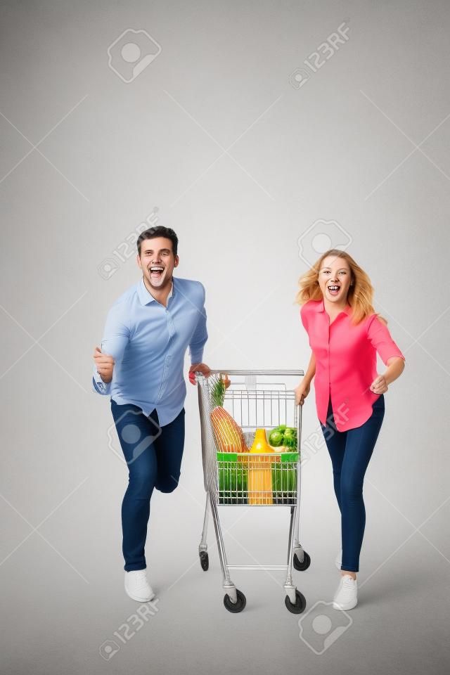 흰색 배경 위에 절연 슈퍼마켓 트롤리와 함께 실행 쾌활 한 커플의 전체 길이 초상화